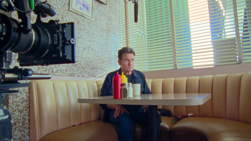 Man sitting alone in a retro diner booth with condiments on the table, film camera and boom microphone visible, sunlight filtering through blinds.