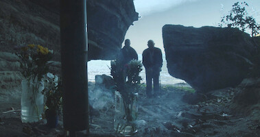 In the foreground, cut flowers and burning incense; in the background two figures visible beyond a rock formation.