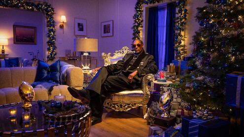 Man in a suit sits on a luxurious chair in an elegant, festively decorated living room with Christmas tree, gifts, candles, and warm lighting, creating a cozy and celebratory mood.