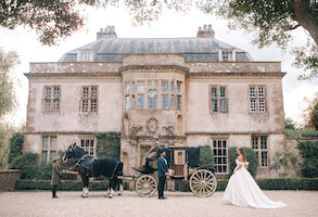 Bride in a white gown stands near a vintage horse-drawn carriage outside an elegant, historic mansion. Lush greenery frames the scene, evoking a romantic, fairytale atmosphere. The mood is dreamy and regal.