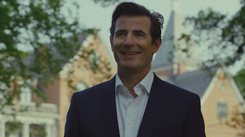 Um homem sorridente usando um traje preto ao ar livre em frente a uma grande construção de tijolos com árvores e uma torre ao fundo, sugerindo um ambiente formal ou acadêmico em um dia ensolarado.