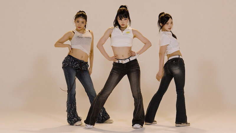 Three women posing confidently in a studio with plain beige background, wearing casual outfits including crop tops, flared jeans, and sneakers, standing in a triangular formation with hands on hips or by their sides.