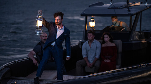 A man in a blue suit holds up a lantern on a small boat at dusk, surrounded by four other people sitting and watching, with a dark sea and another boat in the background.