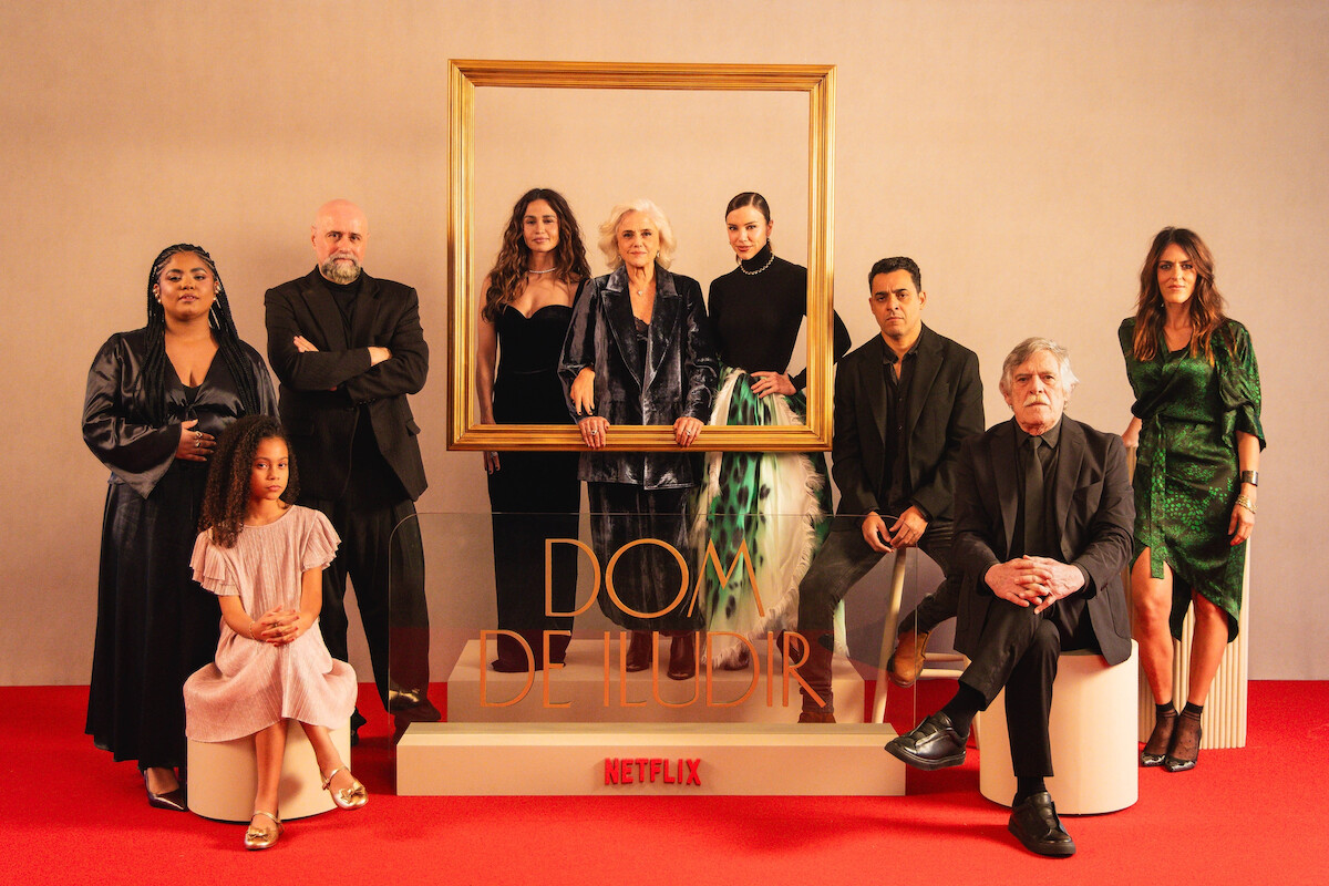 Um grupo de 9 atores da série 'Dom de Iludir' posa em um pequeno palco com o nome da série, sobre chão coberto por um tapete vermelho. As atrizes Nanda Costta, Marieta Severo e Alice Wegmann, no centro da foto, apoiam as mãos em uma grande moldura de quadro dourada