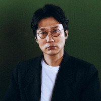 Man with glasses and dark hair wearing a black blazer over a white shirt, sitting against a plain green background in studio lighting.