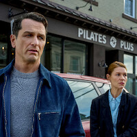 Two serious-looking people stand outside a Pilates studio on a city street, with a car parked behind them; the mood appears tense and contemplative.