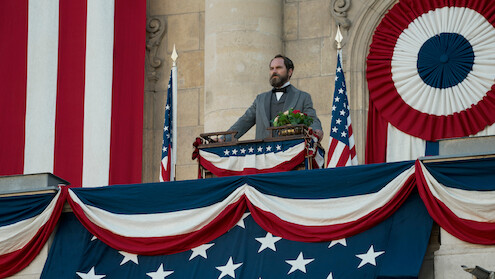 A man in historical attire stands on a decorated balcony with American flags and patriotic bunting, delivering a speech in a formal, historic setting. The mood is solemn and ceremonial.