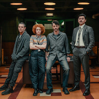 Four people in casual and business attire stand in a dimly lit office with wood paneling, retro décor, tiled floor, and an EXIT sign above a doorway, conveying a detective or investigative setting.