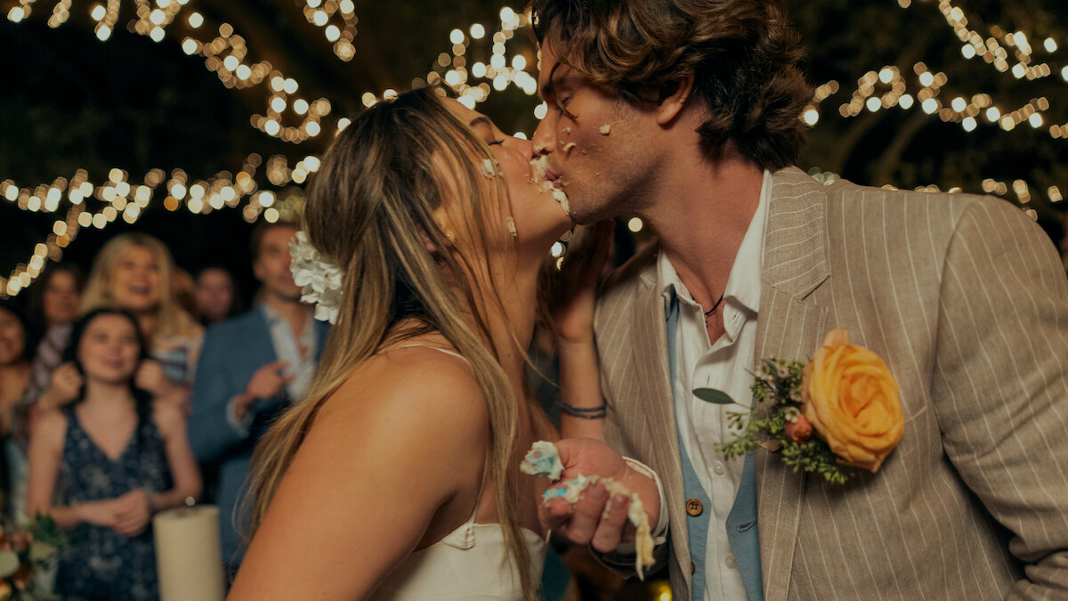 Bride and groom with cake on their faces share a kiss at an outdoor wedding reception. String lights create a festive atmosphere, while guests watch in the background. The groom wears a flower boutonniere.