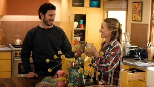 Two people smiling and toasting mugs in a cozy, brightly lit kitchen with yellow cabinets, assorted groceries on the counter, and decorative items in a homey setting.