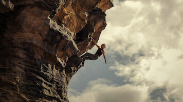 Person rock climbing on a steep, overhanging cliff with dramatic rock formations, reaching outward against a cloudy sky, outdoors in a rugged natural environment.