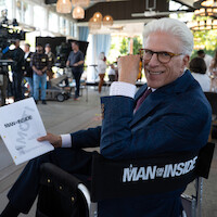 Smiling man in a suit sits on a director’s chair labeled "Man on the Inside," holding a script on a bright, busy film set with crew and equipment in the background.