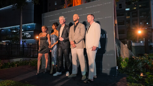 Five well-dressed people posing together on an outdoor evening event stage, city buildings and greenery in the background, creating a formal and celebratory mood.