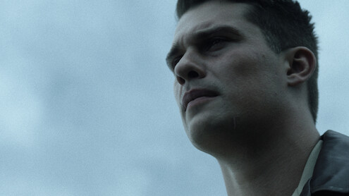 Young man with short dark hair in a serious expression, shown in close-up against a cloudy sky, outdoors in natural light.