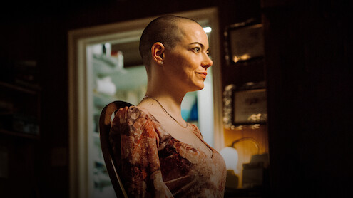 A woman with a shaved head sits in profile in a warmly lit room, smiling. The background includes a doorway revealing a bright, cluttered space and certificates on the wall, suggesting a personal or home environment.