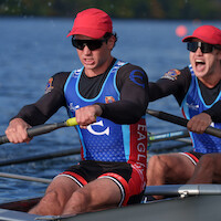 Two male rowers in sunglasses and matching athletic gear intensely rowing a boat on a lake during a race, with blue water, blurred buoys, and trees in the background, expressing focus and teamwork.