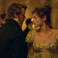 A man and woman in historical attire stand close together at a dimly lit, elegant ballroom event, with the woman in a floral dress and the background softly blurred, creating a romantic atmosphere.