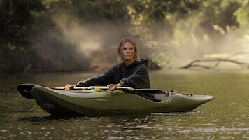 A person wearing a dark hoodie is paddling a green kayak on a calm river, surrounded by trees with mist rising in the background, creating a serene outdoor nature scene.