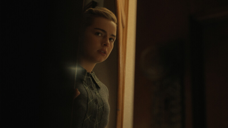 Addison Rae with a concerned expression peeks around a doorway into a dimly lit room.
