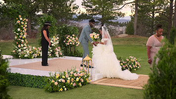 Outdoor wedding scene with bride and groom at altar, surrounded by flower arrangements and greenery, officiant present, another person walking away, trees and mountains in the background, peaceful and romantic setting.