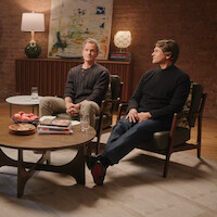 Two men sit in armchairs in a cozy living room with a brick wall, abstract art, and lamps behind them. In front, a coffee table holds books, snacks, and glasses of water, creating a relaxed, conversational setting.