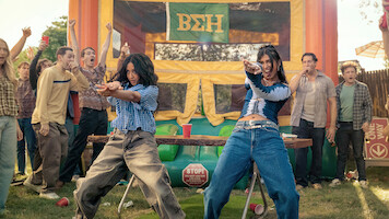 Two women dance energetically at an outdoor party with a crowd of cheering people, in front of a colorful bouncy house decorated with Greek letters, on a sunny day.