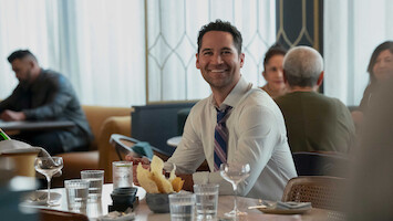 Smiling man in dress shirt and tie sitting at a restaurant table with chips, water glasses, and wine glasses, with other people dining in the background. Bright, modern interior with natural light.
