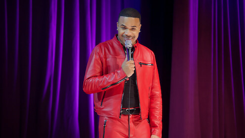 A comedian in a bright red leather outfit performs stand-up on stage, holding a microphone, with purple stage curtains in the background and stage lighting.