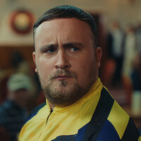 A man with short hair and a trimmed beard wearing a yellow and blue sports jacket is indoors, with blurred people and warm lighting in the background, suggesting a social or competitive event setting.