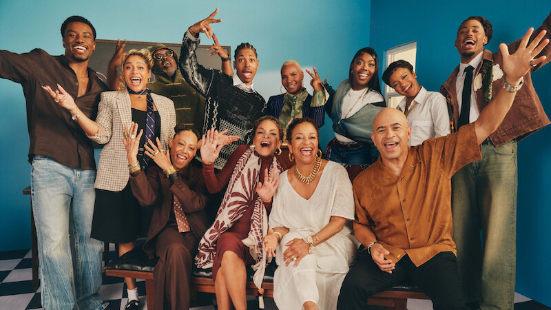 Group of people in stylish clothing smiling and posing energetically in a brightly lit room with blue walls and checkered floor, conveying a sense of joy and celebration.