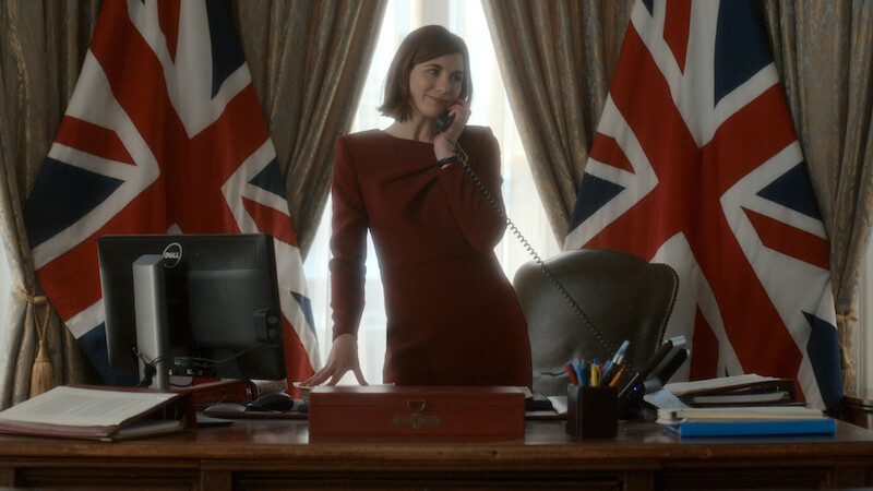 A woman in formal attire stands behind a large desk, speaking on a phone in an office decorated with two large UK flags, books, a computer, and office supplies, suggesting a government or official setting.