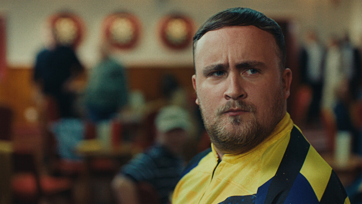 A man in a yellow and blue sports shirt looks serious in a lively indoor setting with people and dartboards in the background, suggesting a darts competition or social club atmosphere.