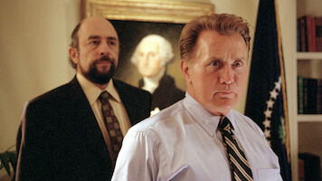 Two men in business attire stand in an office with a U.S. flag and a portrait of George Washington in the background, suggesting a formal or political setting.