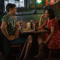 A man and woman sit at a table in a cozy restaurant at night, smiling and talking over drinks and food, creating a warm, intimate atmosphere.