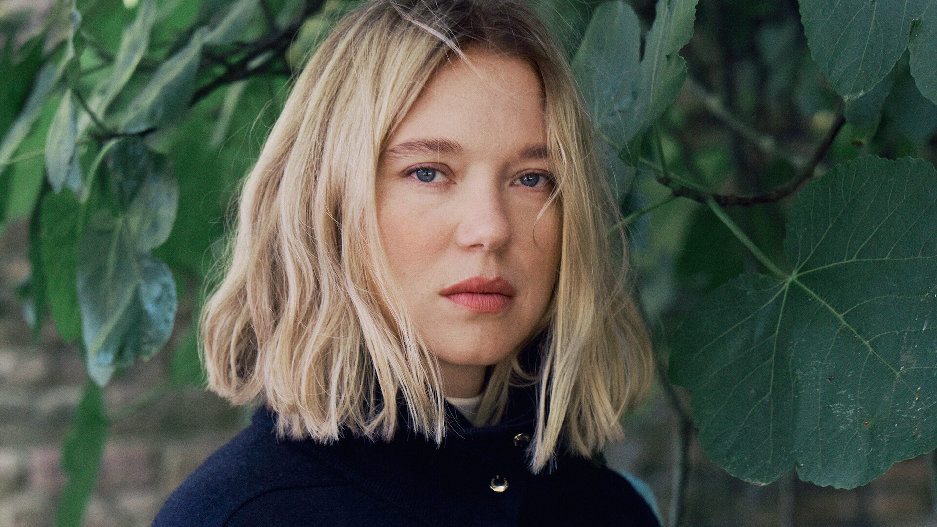 A woman with shoulder-length blonde hair stands outdoors in front of large green leaves and foliage, wearing a dark coat and looking at the camera with a neutral expression.
