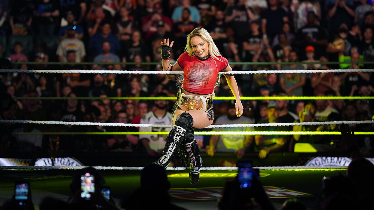 A female wrestler wearing a championship belt poses confidently in a wrestling ring, with a cheering crowd and bright arena lights creating an energetic and celebratory atmosphere. Fans are capturing the moment on their phones.