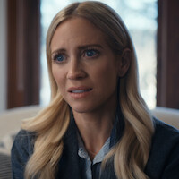 A concerned woman with long blonde hair sits indoors by a window in a softly lit room, wearing a blue dress shirt and sweater, looking slightly distressed or worried.