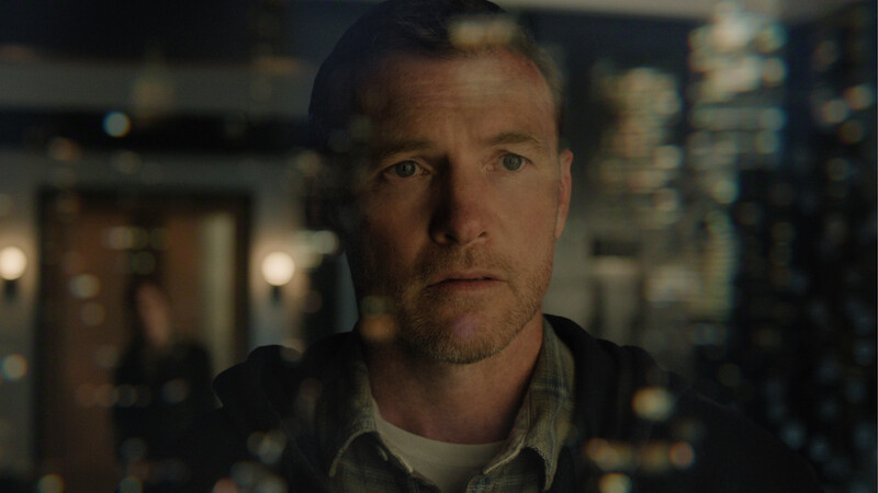 A man stands indoors, looking intently through a glass wall with light reflections; the environment is dimly lit with modern decor, and a blurred figure is visible in the background.