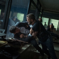 Two men struggle violently in an office, one pinning the other against a desk, while others watch in shock. The setting is a cluttered workspace with glass walls and scattered files, lit by daylight through window blinds.