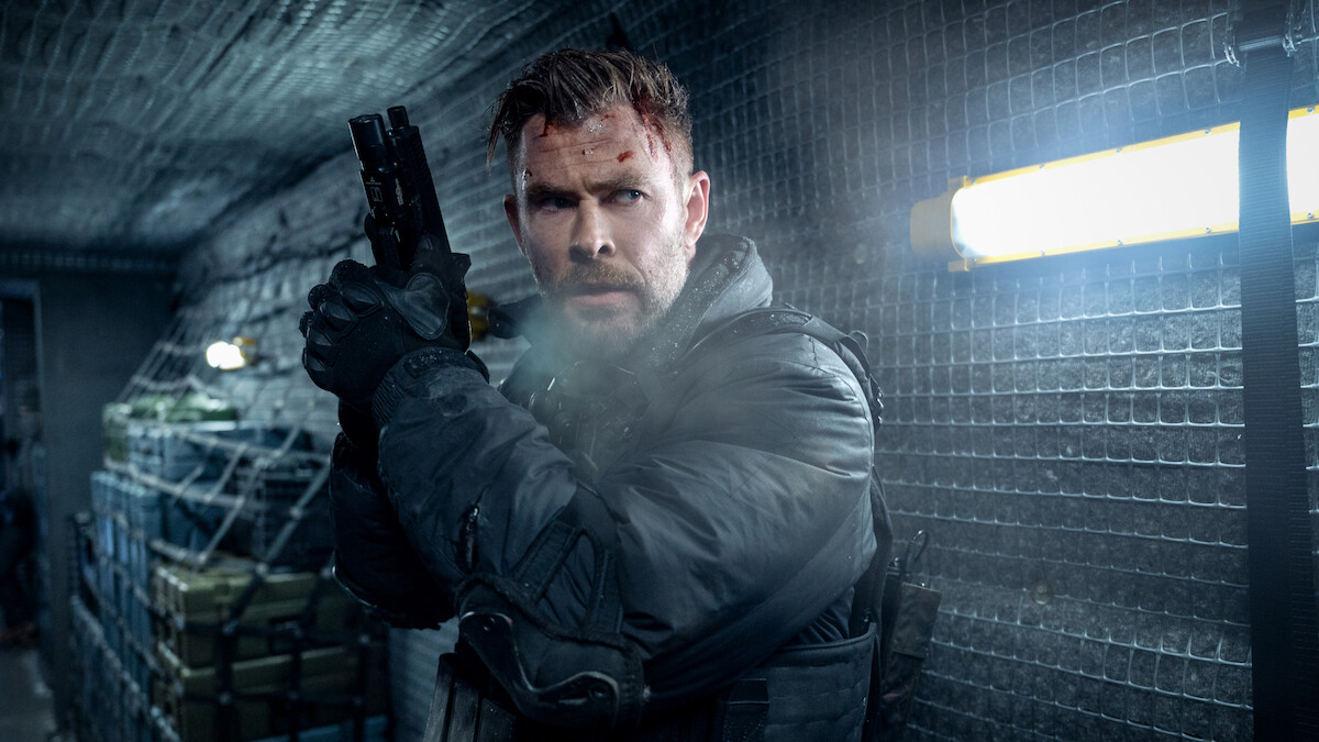 Chris Hemsworth as Tyler Rake with a bruised face and tactical gear holding a handgun, standing alert in a dimly lit, metal-walled corridor with supplies and fluorescent lighting, appearing ready for action or conflict.