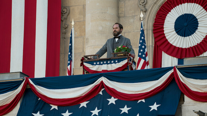 A man in historical attire stands on a decorated balcony with American flags and patriotic bunting, delivering a speech in a formal, historic setting. The mood is solemn and ceremonial.