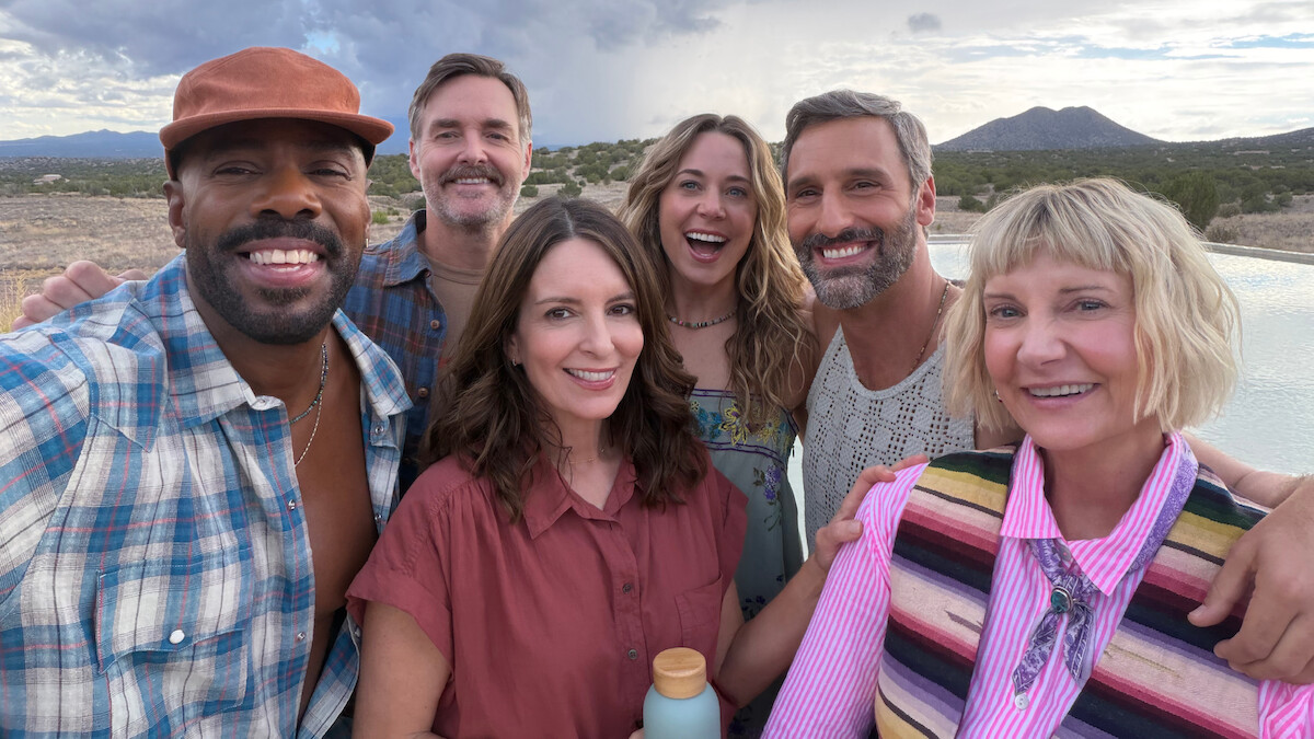 Group of six smiling adults taking a selfie outdoors by a pool, with mountains and cloudy sky in the background, casual clothing, relaxed atmosphere, desert landscape.