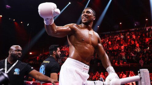 A victorious boxer with white gloves and shorts smiles in a brightly lit boxing ring, surrounded by a cheering crowd and security staff, creating an energetic and triumphant atmosphere.