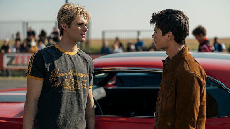 Two young men stand facing each other next to a red car at an outdoor event, with spectators and a fence visible in the background on a sunny day.