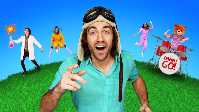 Danny GO! in an aviator hat smiling on grass under blue sky, with scientist holding beaker, person with pink bats, woman jumping, and bear costume playing drums labeled "Danny Go!" in colorful, playful setting.