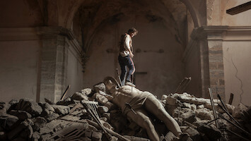 A person standing on rubble inside a damaged, old stone building, with a large fallen classical statue lying among the debris under an arched ceiling, evoking a sense of destruction and loss.