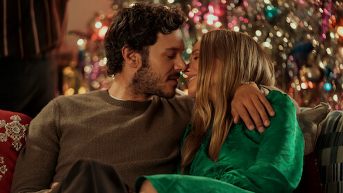 A couple sitting close together on a couch, gazing lovingly at each other, with a decorated Christmas tree glowing in the background, creating a warm, festive indoor holiday atmosphere.