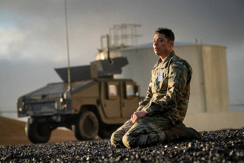 Um soldado de uniforme camuflado se ajoelha em um terreno rochoso perto de um veículo militar. Ao fundo, um tanque industrial aparece sob o céu nublado, em uma imagem de clima contemplativo e solene.