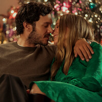 A couple sits closely together, smiling and about to kiss, in front of a decorated Christmas tree with colorful lights, creating a warm and festive holiday atmosphere.