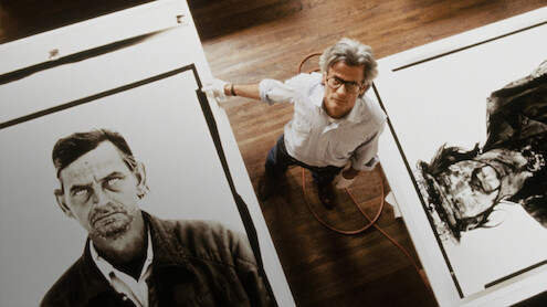 Man standing in studio on wooden floor, surrounded by large black-and-white portrait artworks on paper, looking up at the camera.